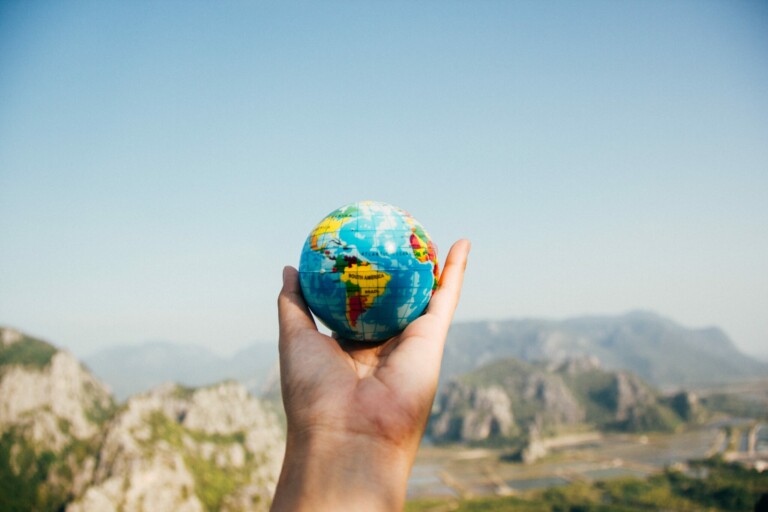 A hand is holding a small model of a globe in an outdoor setting, symbolizing international tax minimization strategies. The background features a clear blue sky and distant mountains with green vegetation.