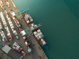 Aerial view of a busy port in Costa Rica showing multiple shipping containers neatly stacked along the dock and large cargo cranes unloading a cargo ship docked at the pier, bustling with exports. The water adjacent to the port is calm and turquoise.