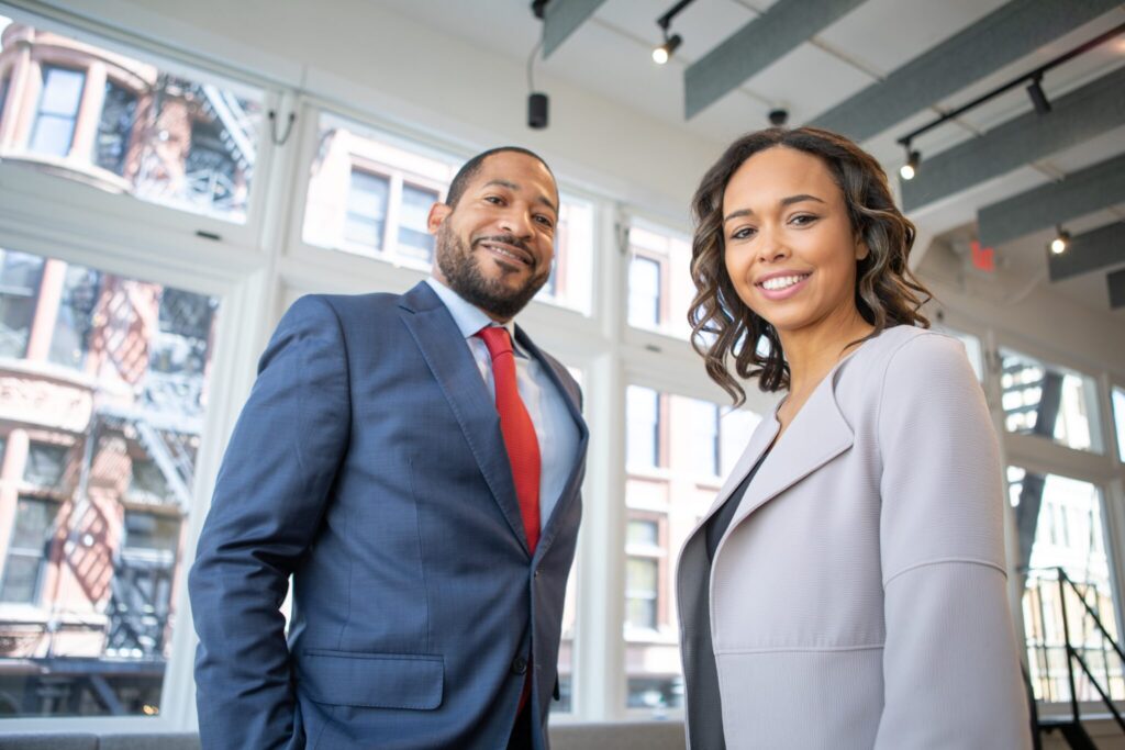 Two people standing indoors in a bright room with large windows and an urban view. They are dressed in business attire; the man is wearing a blue suit with a red tie, and the woman is in a light gray blazer. Both are smiling and looking towards the camera, ready to do business in El Salvador.