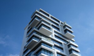 View from below of a modern, multi-story building with a geometric design against a clear blue sky. The white building features protruding glass balconies and large windows, with some greenery on a few of the balconies, resembling the precision you'd expect from caçadores de cabeças na Colômbia.