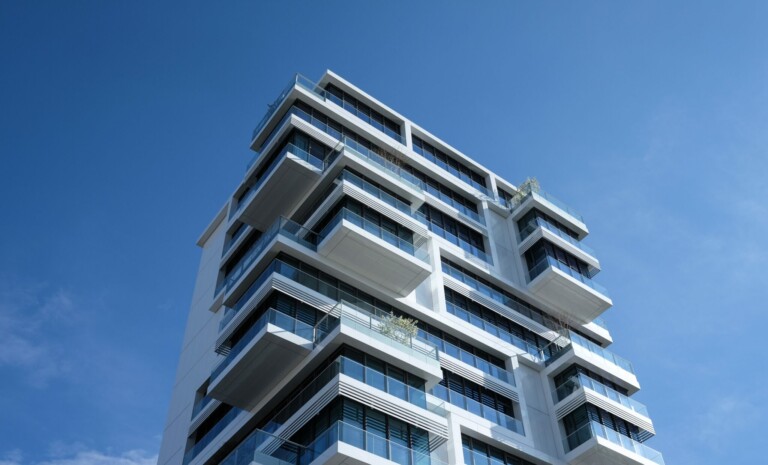 View from below of a modern, multi-story building with a geometric design against a clear blue sky. The white building features protruding glass balconies and large windows, with some greenery on a few of the balconies, resembling the precision you'd expect from caçadores de cabeças na Colômbia.