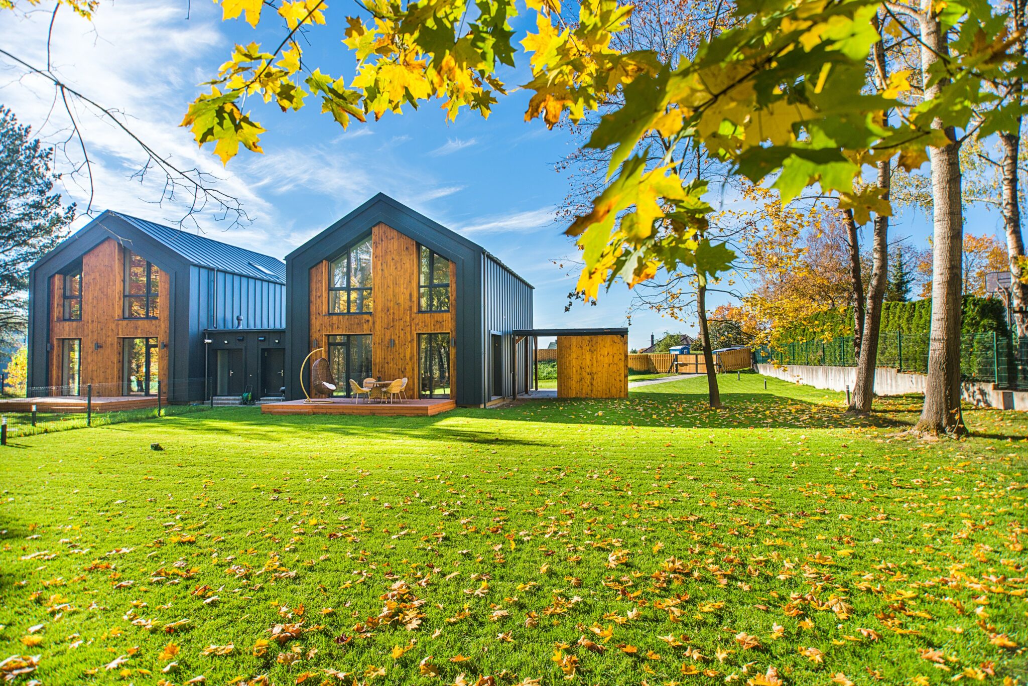 A modern two-story house with large windows and wooden exterior panels, set in a spacious yard with green grass and scattered fallen leaves. Trees with autumn foliage surround the property under a blue sky—your ideal opportunity to buy property in Chile.