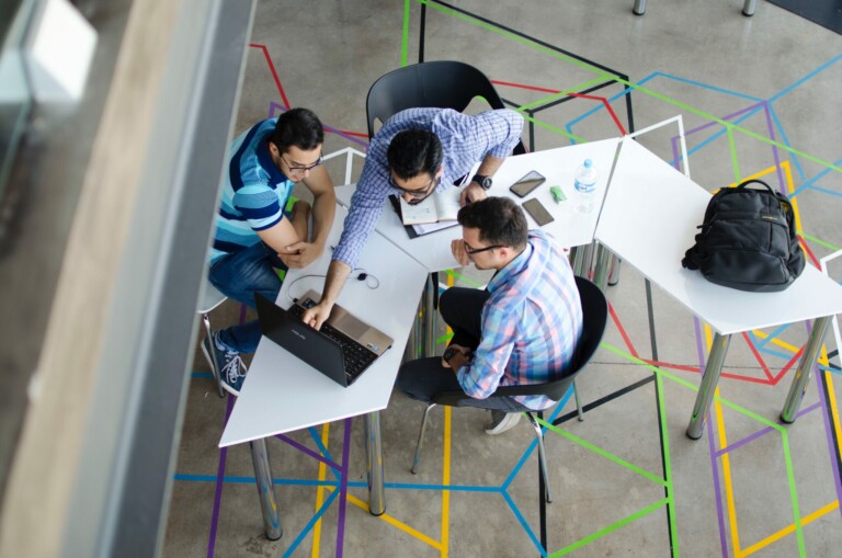 Three people sit at white desks arranged in a U-shape, working together with laptops and documents, possibly planning to form an NGO in El Salvador. The office floor has a colorful geometric pattern design. A backpack and a water bottle are on one of the desks. The atmosphere appears collaborative and industrious.