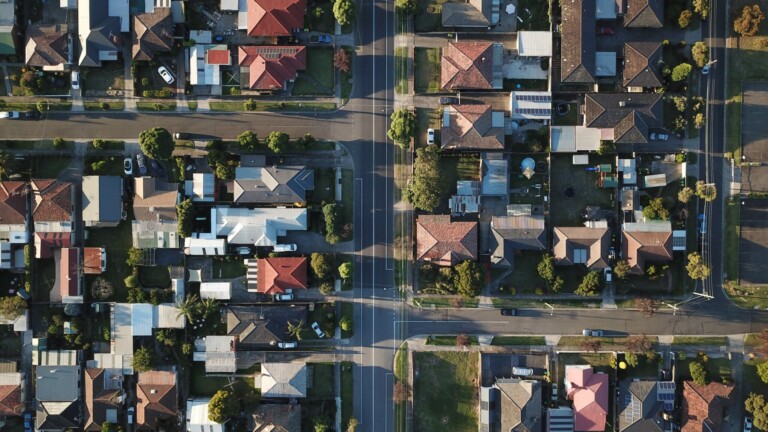 Aerial view of a residential neighborhood featuring a grid of houses with various roof shapes and colors. Streets separate the blocks of houses, with cars parked along the curbs. Trees and yards are visible throughout the area, creating a suburban environment reminiscent of buying property in Bolivia.