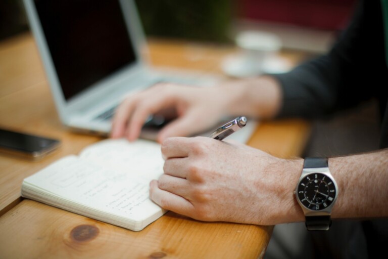 A person wearing a watch is writing in a notebook with a pen. Their other hand is resting on an open laptop on a wooden table, perhaps organizing an Entity Health Check in Panama. A smartphone is placed to the side of the notebook, and a blurred cup is visible in the background.