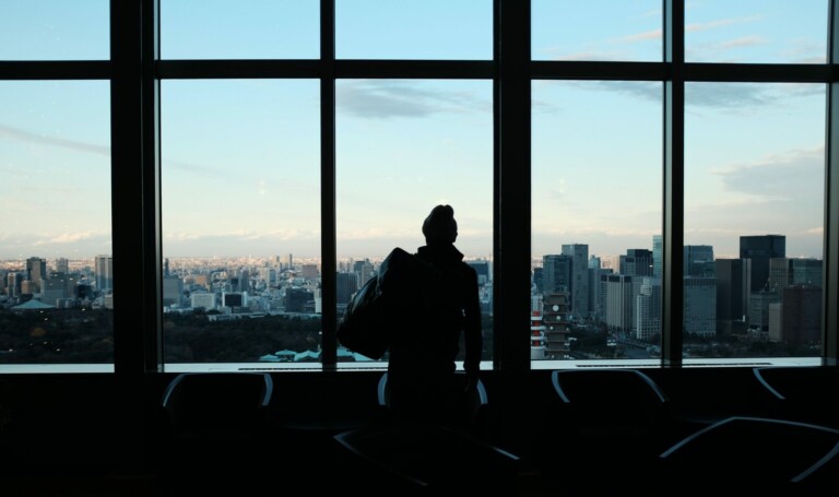 Una silueta de una persona se encuentra frente a ventanas del piso al techo, con vistas a un paisaje urbano con numerosos edificios de gran altura bajo un cielo despejado. La escena, posiblemente tomada desde una oficina interior perteneciente a una PEO en Perú, presenta varias sillas alrededor del individuo.