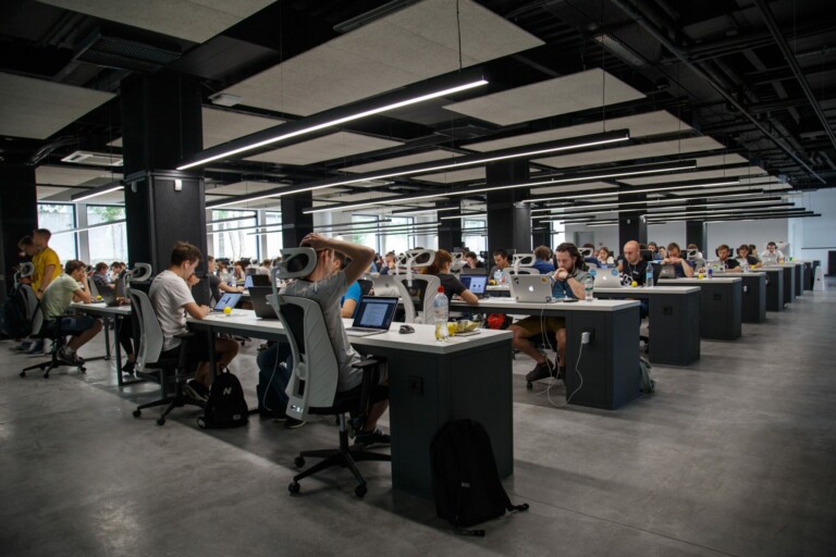 A large office space in Peru features rows of people working at desks with computers. The room boasts a modern design with black ceiling fixtures, ample natural light from large windows, and a concrete floor. Backpacks and personal items are placed by some desks, enhancing connectivity for business in the area.