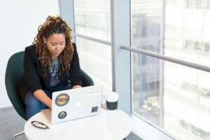 A person with long, curly hair sits in a modern office space, typing on a laptop adorned with various stickers. They are brainstorming ideas to form an NGO in Argentina. Seated in a green chair by a window with a cityscape view, they have a coffee cup and small dish on the round white table before them.