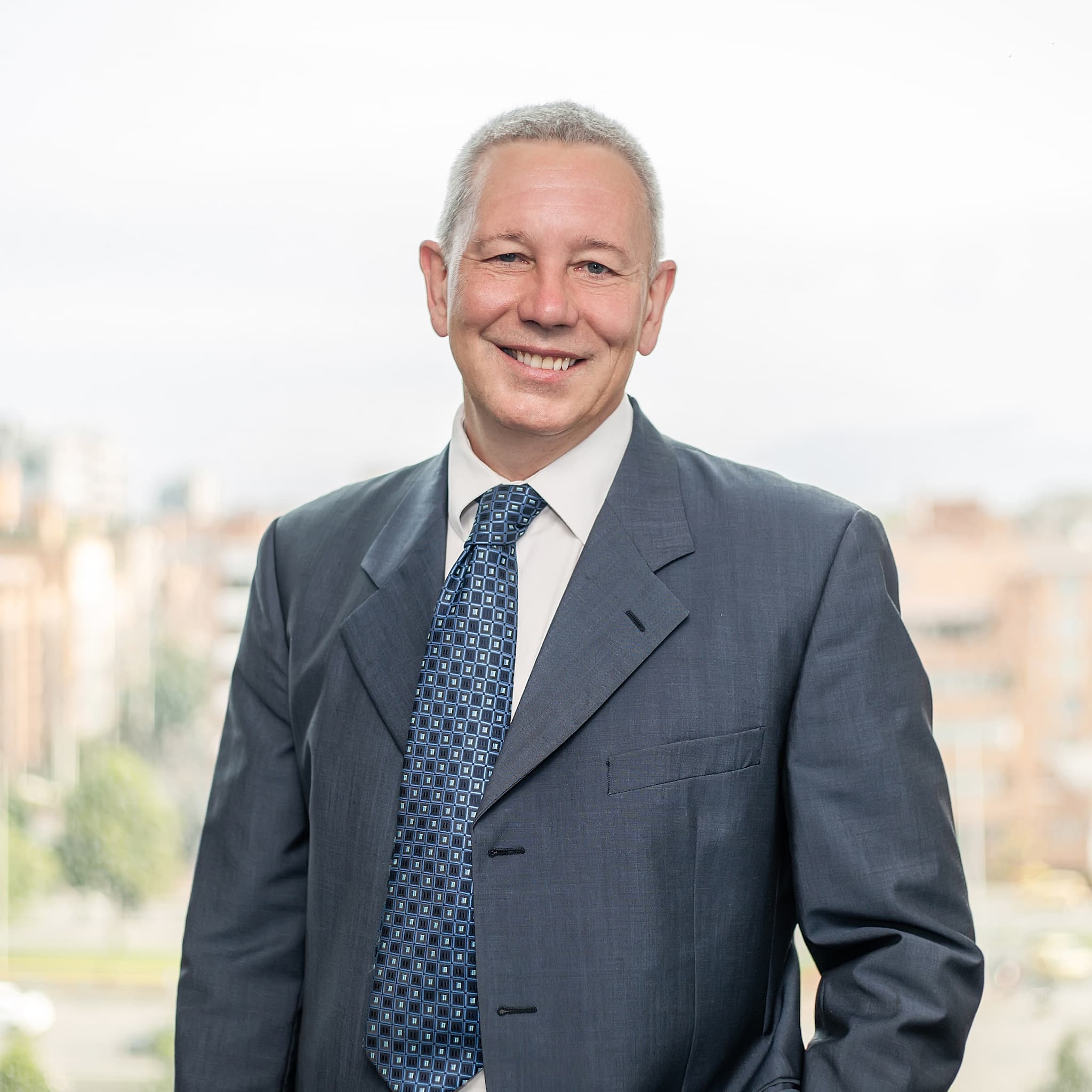 David Wright in a grey suit and blue tie smiling, standing against a blurred cityscape background.