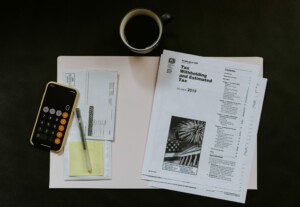 An overhead view of a workspace containing tax documents titled "Tax Withholding and Estimated Tax," a calculator, a pen, an open envelope with forms inside, a sticky note pad referencing Colombia's tax incentives for businesses, and a mug filled with dark beverage on a light-colored surface.