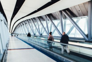 A modern pedestrian walkway features a ceiling with a wavy design and large structural beams. Several people, including a man in a suit possibly on his way to secure a Business Visa in Guatemala and a woman with a bun, walk in opposite directions, separated by a glass partition in the center.