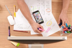 A person is working at a desk with design sketches, an iPhone, colored markers, a mouse, and a computer monitor. Amidst the scattered writing tools, they're holding a pink highlighter and marking a paper while contemplating an upcoming Entity Health Check in Panama.