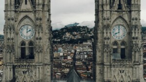 Two tall stone clock towers with blue dials showing just after 9:15 and 3:15, framing a view of a city with a mountain in the background. The towers feature gothic architecture, much like how Ecuador's double taxation agreements bring historical context to modern financial landscapes.