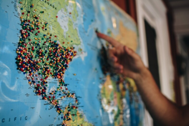 A hand is pointing at a specific location on a world map filled with numerous colorful pushpins, particularly dense in North America. The background is slightly out of focus, and the map includes visible country names and geographic details, highlighting import and export regulations in Belize.