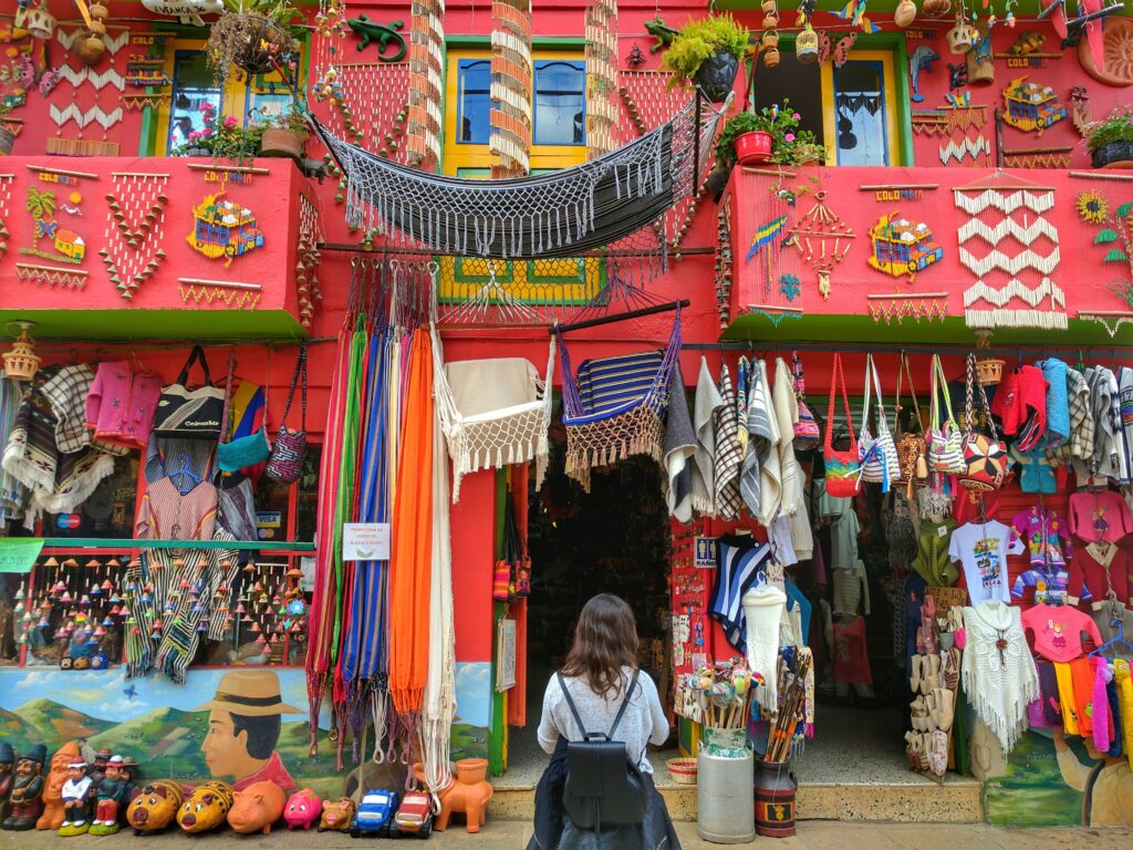 A person with a backpack stands in front of a vibrant, colorful market stall. The stall is adorned with textiles, woven items, clothing, and various handicrafts—a reflection of the rich cultural tapestry and business etiquette in Colombia. The walls of the building are painted red, and the stall is overflowing with goods.