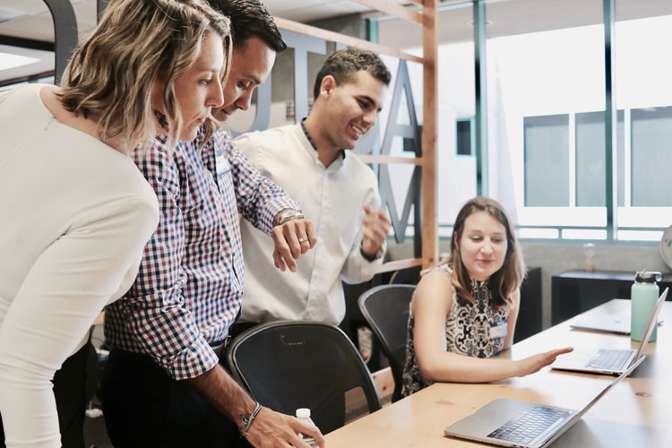 Four people are gathered around a desk in an office. Three are standing, looking at a laptop, while one person is seated and typing. The seated person is wearing a sleeveless dress, and the others are casually dressed. They appear to be collaborating on payroll outsourcing in Chile.