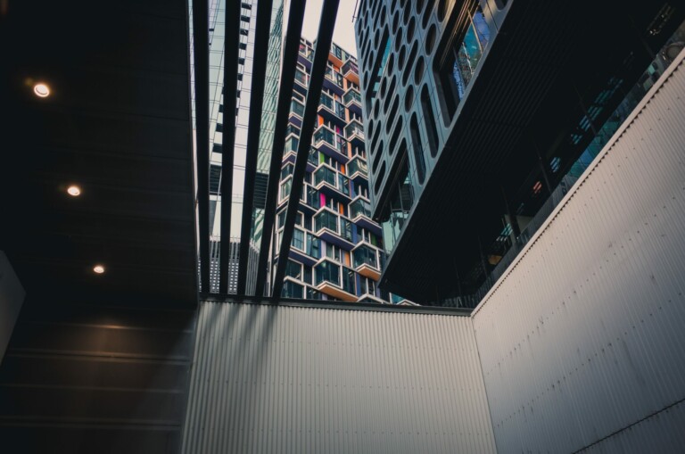 View of modern urban architecture from a low angle, showcasing multiple buildings with geometric designs and colorful balconies framed between intersecting overhead structures. Overhead lighting fixtures are visible on the left side, much like a company formation agent in the Dominican Republic guides new ventures.