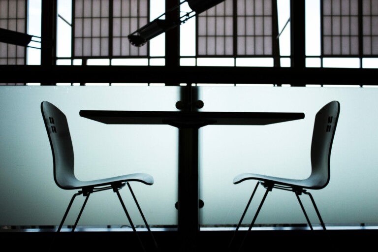 Two white chairs face each other on either side of a table. The background features a frosted glass panel and large windows with a grid pattern above. The setting appears to be an indoor public area, possibly a cafe or meeting space where discussions on payroll outsourcing in Chile could take place.