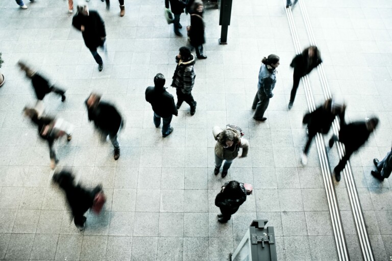 A high-angle view of people walking in different directions on a tiled floor. A lawyer in Uruguay is among the crowd, creating a vivid contrast between motion and stillness. The scene appears busy with a mix of walking and stationary people, some figures blurred by their movement.