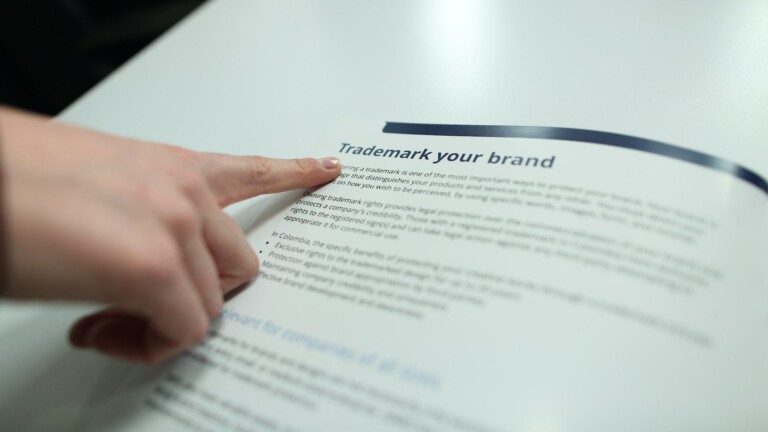 A person's hand points to content on a printed document titled "Trademark Your Brand." The text below the title discusses the importance and benefits of trademarking a brand, though the specific details are not fully legible. The document rests on a white surface, possibly observed by caçadores de cabeças na Colômbia.