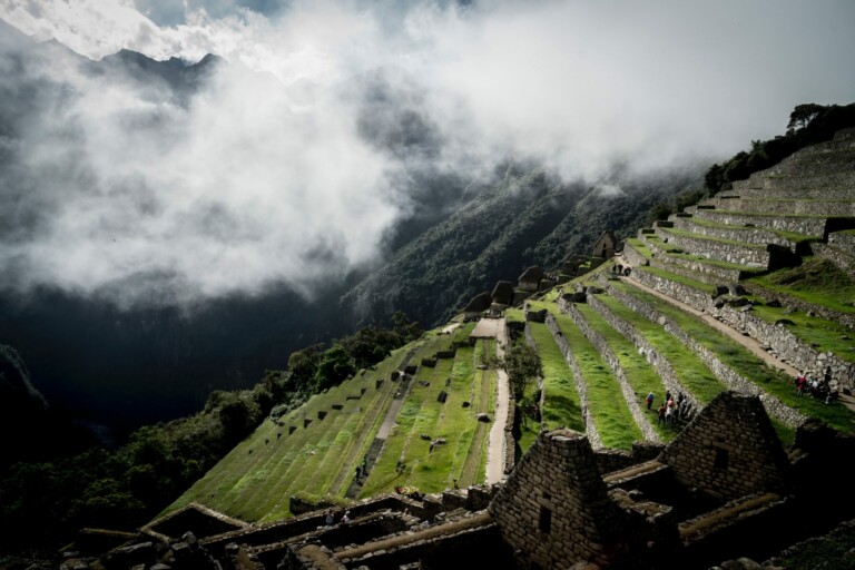 A misty view of terraced fields and ancient stone structures at Machu Picchu. The background consists of cloudy, forested mountains. Amid various visitors exploring the Incan archaeological site, whispers of corporate restructuring in Peru hint at a changing landscape beyond these historic terraces.