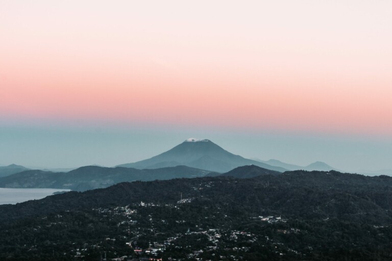 A distant cone-shaped mountain surrounded by lower, hilly terrain rises majestically. The sky is a gradient, transitioning from a soft pink near the horizon to light blue higher up. In the foreground, densely forested areas with scattered buildings set the scene, reminiscent of where you might find a company formation agent in El Salvador.