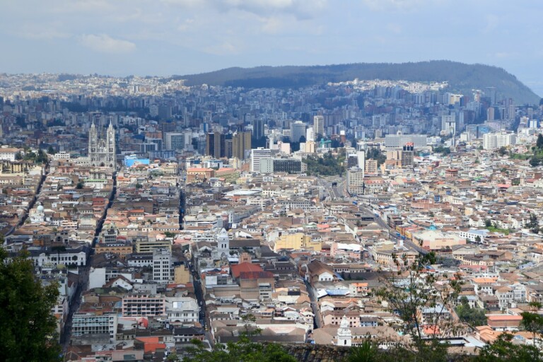 Aerial view of a densely populated city featuring a mix of modern and historic buildings. The image captures a sprawling urban landscape with varied architecture set against green hills and a partly cloudy sky—an inspiring scene for anyone considering starting a business in Ecuador.