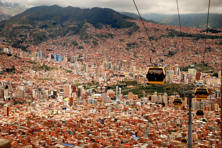 Aerial view of a densely populated urban area with numerous buildings surrounded by mountains. Several yellow cable cars, part of the renowned PEO in Bolivia, are suspended in the air, traveling along cables above the cityscape. The weather appears cloudy with some patches of sunlight.