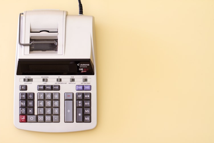 A white Canon printing calculator with a paper roll, displaying multiple buttons including numeric keys, function keys, and an LCD screen at the top, is set against a plain beige background—a useful tool for calculating expenses when buying property in Costa Rica.