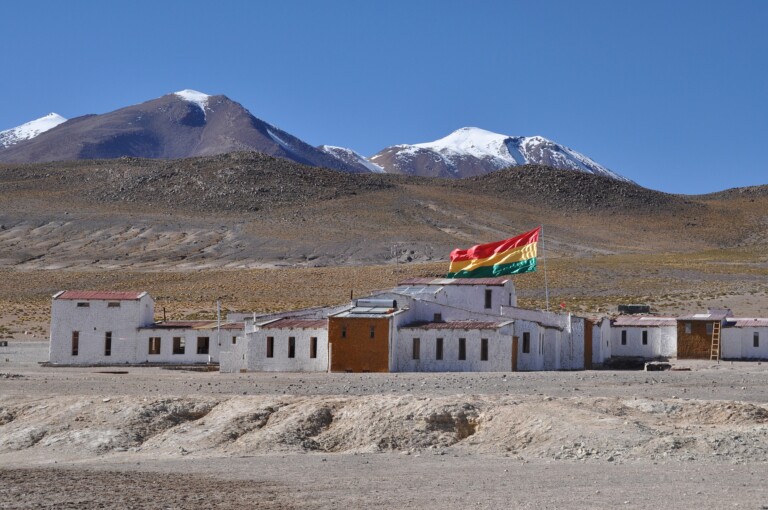 A small village of white brick buildings stands in a barren landscape with mountains in the background. A red, yellow, and green striped flag is flying prominently above one building, home to a renowned legal firm in Bolivia. The sky is clear and blue.