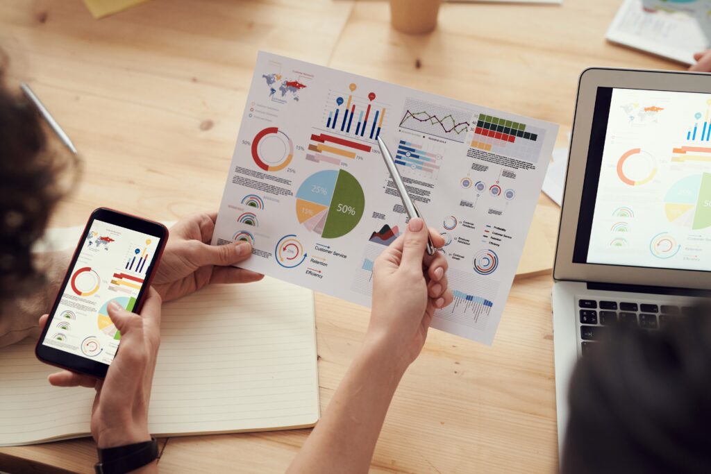 Three people are analyzing colorful charts and graphs displayed on a printed sheet. One person is pointing at the sheet with a pen, another is holding a smartphone displaying similar data, and a laptop showing charts for an Entity Health Check in Uruguay is partially visible.