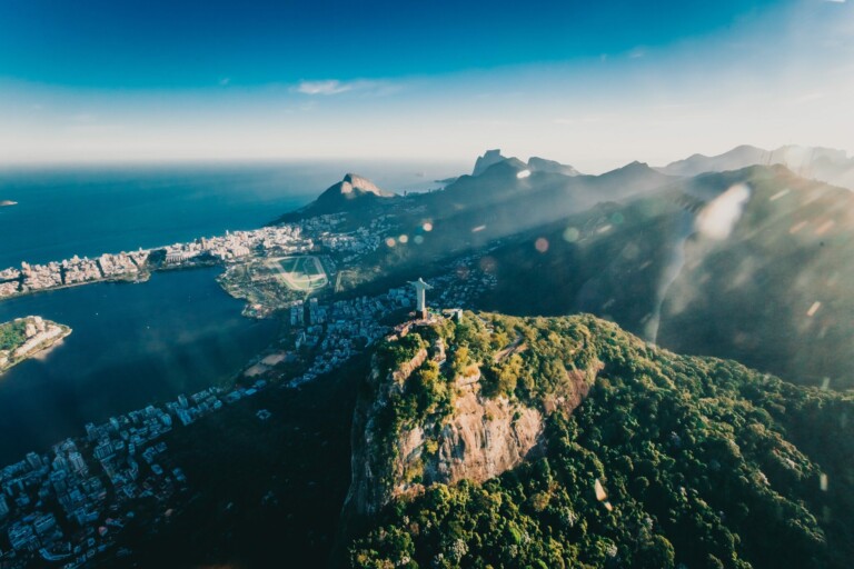 Aerial view of Rio de Janeiro with the iconic Christ the Redeemer statue on a mountain in the foreground. The vibrant cityscape, surrounding hills, coastline, and ocean are visible under a clear blue sky. The image captures both natural beauty and urban development of Brazil's bustling startup hub.