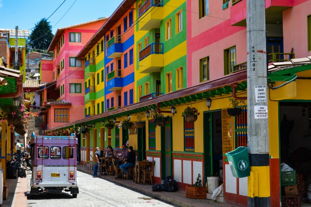 A colorful street with buildings painted in bright pink, green, yellow, and blue. The street features outdoor seating areas where people are sitting and talking, perhaps discussing the latest from tech companies in Colombia. A vehicle is parked on the left side of the street. A utility pole with signs is visible on the right.