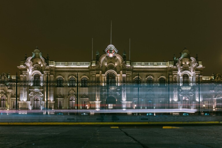 A nighttime photograph of an ornate, illuminated building with an intricate facade and multiple arched windows. The building, which once hosted prominent tech companies in Peru, stands behind a black metal fence. Light streaks from passing vehicles are visible in the foreground.