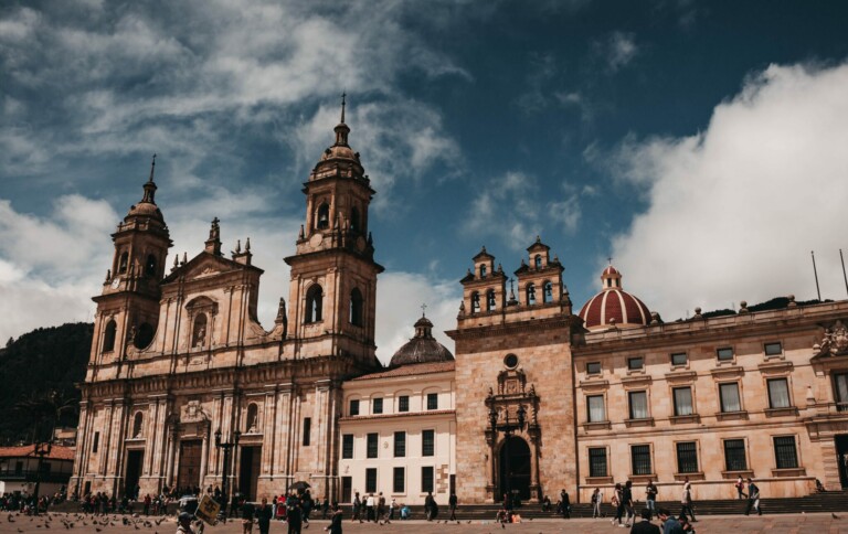 A historic cathedral and adjacent buildings stand under a partly cloudy sky. The structures feature ornate architectural details and towers. People—among them, visitors seeking Corporate Legal Counsel in Colombia—gather at the base, some walking while others stand. Pigeons can be seen in the foreground.