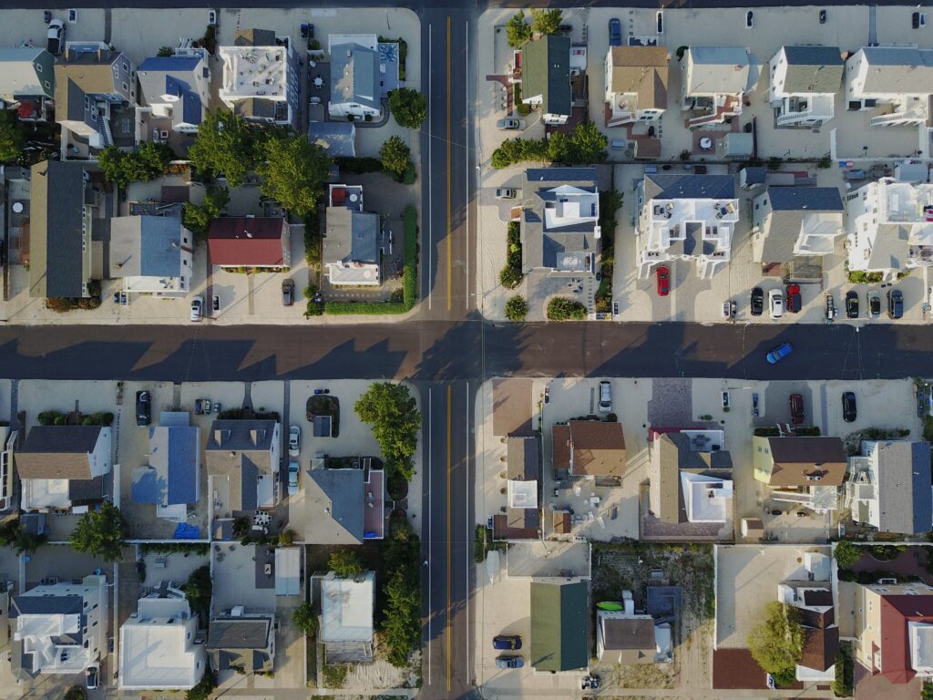 Aerial view of a residential neighborhood featuring two intersecting roads. The houses are arranged in neat rows, with some greenery and parked vehicles visible. Shadows of the buildings stretch across the streets in daylight, showcasing an ideal setting for buying property in Costa Rica.