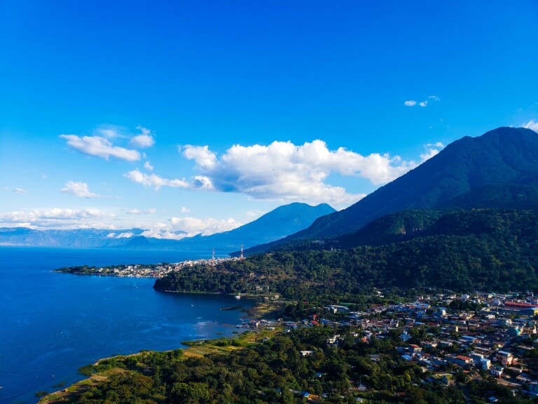 Aerial view of a coastal town along the edge of a large lake with lush green hills and mountains in the background. The town has numerous buildings, while the lake appears calm under a bright blue sky with scattered clouds, reminiscent of the serene landscapes far removed from caçadores de cabeças na Colômbia.