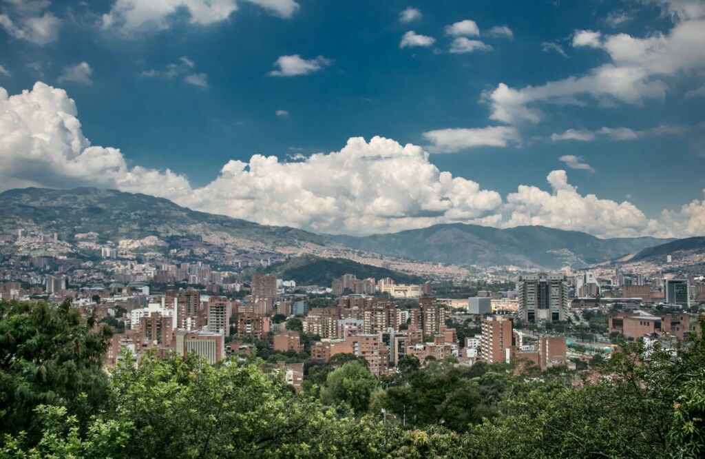 A panoramic view of a city with a mix of high-rise buildings and lower structures surrounded by greenery. Mountains are visible in the background under a partly cloudy sky, highlighting the vibrant landscape where numerous tech companies in Colombia innovate and thrive.