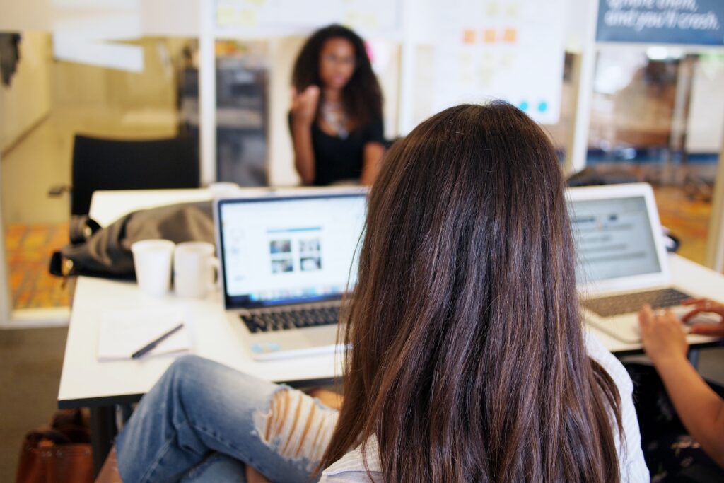 A person with long brown hair sits at a table with a laptop, facing another blurred individual in the background. The table also holds notebooks, a pen, two white mugs, and another person using a laptop. The setting appears to be an office or meeting room of one of the tech companies in Peru.