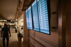 A series of digital flight information display screens at an airport show arrival times. The screens are positioned on a wall, and an individual holding a beverage walks by in the corridor. In the background, other travelers and airport signage provide information about obtaining a Business Visa in Brazil.