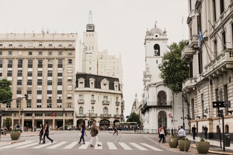 People walking across a crosswalk at a busy intersection with historic buildings in the background, including a white bell tower. Traffic lights and buses are visible, along with large planters on the sides of the street—an everyday scene reminiscent of EOR na Argentina.