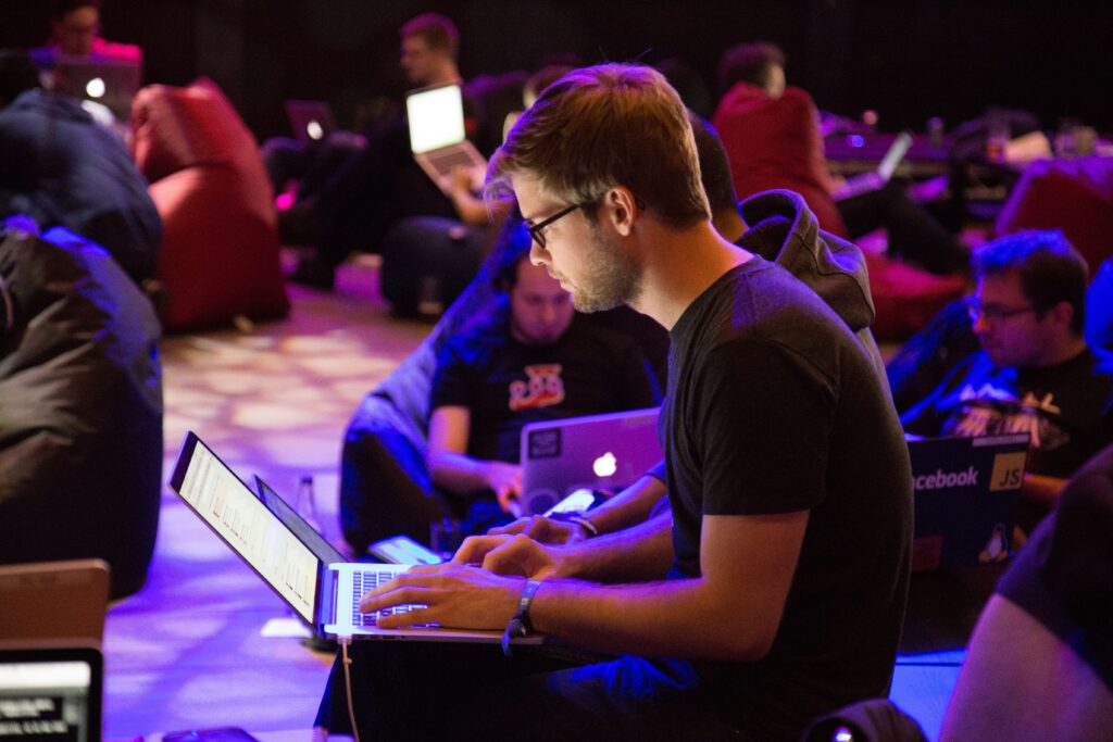 A group of people are seated on bean bags and chairs working on their laptops in a dimly lit room. The focus is on one person in the foreground wearing glasses and a dark t-shirt, typing on a laptop with a white screen. The scene reflects the vibrant work culture of tech companies in Colombia.