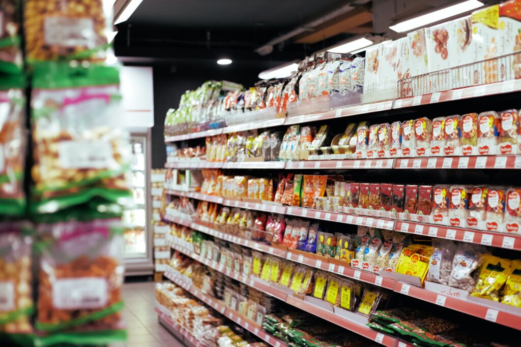 A grocery store aisle displays a variety of packaged food items. Shelves are stocked with an assortment of snacks, cereals, and other dry goods. The left side shows hanging bags of snacks, each adhering to product registration standards in Chile. The background includes refrigeration units.