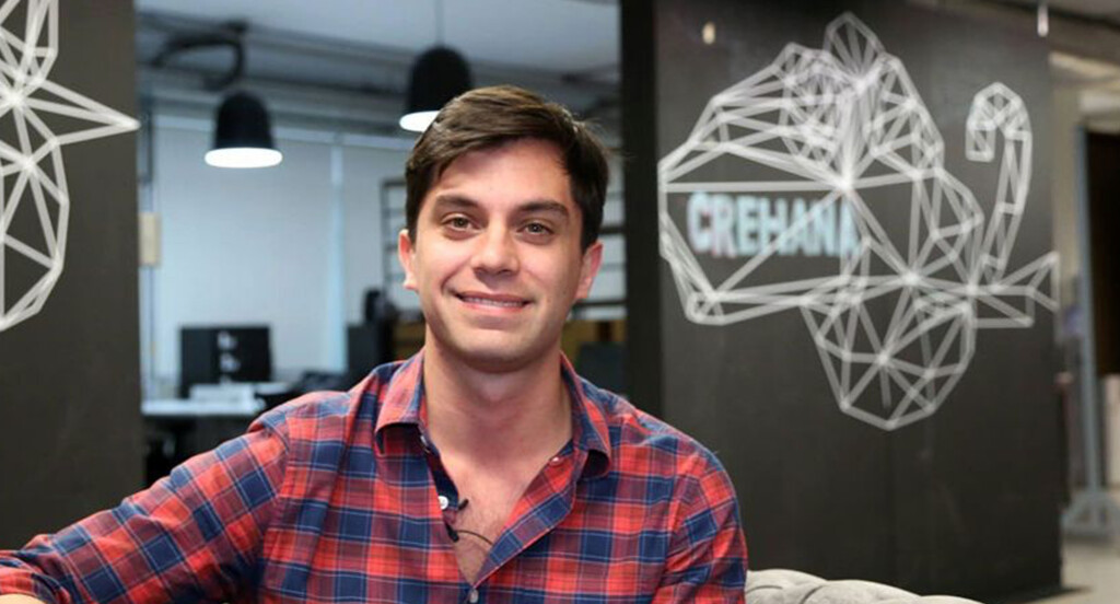 A man with short dark hair and wearing a red and blue plaid shirt is smiling and looking at the camera. Behind him is a black wall featuring a geometric animal design and the word "CREHANA" in white text, representing one of the prominent tech companies in Peru. The background includes various office furnishings.