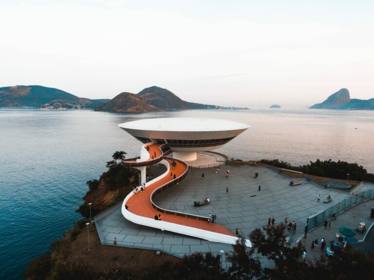 Aerial view of the Niterói Contemporary Art Museum in Brazil, featuring its distinctive saucer-shaped structure on a cliffside overlooking the water. A winding red walkway leads to the entrance, with people walking around the museum's exterior. Mountains can be seen in the background, embodying the rich culture supported by many an ONG no Brasil.