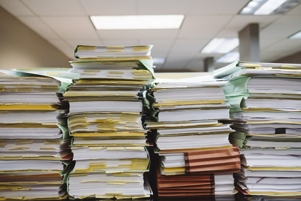 Stacks of paperwork and files for the Entity Health Check in Uruguay are arranged on a desk in an office setting. The folders, in various colors including green and brown, are filled with a large number of papers. Office lighting is visible above.