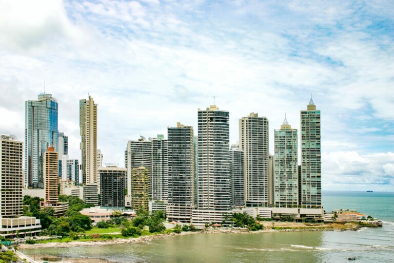 A coastal cityscape featuring numerous modern high-rise buildings on the shoreline, reflective of Panama's booming tech companies. The buildings vary in height and design, with a mix of residential and commercial structures. The sea is visible to the right under a partly cloudy sky.