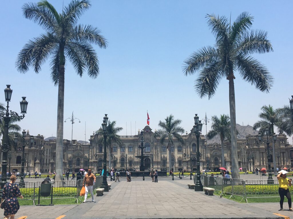 A large historic building with ornate details stands under a clear blue sky. Palm trees are in the foreground, and people walk on the paved plaza and surrounding grassy areas. A flag is seen on top of the building, symbolizing pride much like the burgeoning tech companies in Peru. The setting appears to be a public square.