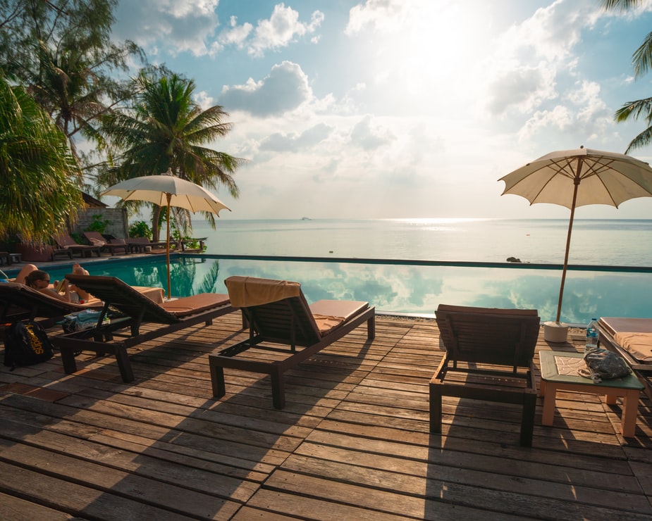 Outdoor pool area with several lounge chairs facing a calm sea. Two umbrellas provide shade, tall palm trees are visible, and the sky is partly cloudy with the sun shining. The serene scene suggests a relaxing, tropical setting, perfect for unwinding after meetings with tech companies in Peru.