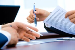 Two people are sitting at a table, each holding pens and engaged in a discussion. One person, talking about caçadores de cabeças na Colômbia, is pointing at a document with text, which the other person is holding. A laptop is partially visible in the background.